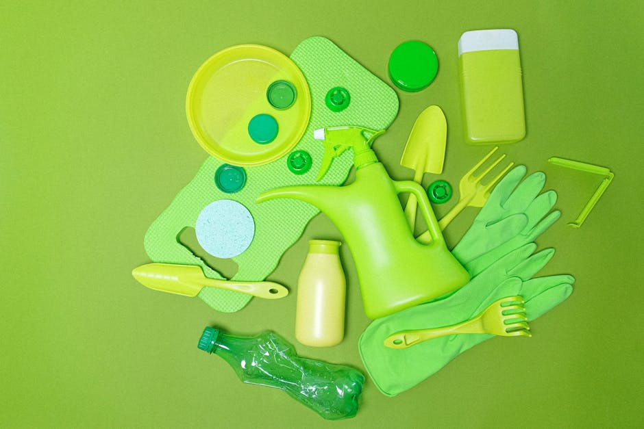 A collection of gardening tools and supplies arranged on a green surface, including a bright yellow watering can, a set of green rubber gloves, a small yellow trowel, a yellow rake, a green spray bott